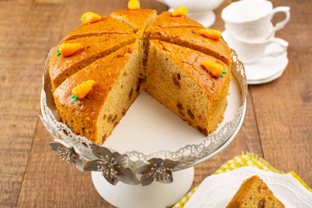 A beautifully arranged carrot cake on a cake stand, sliced and decorated with tiny sugar carrots, with a white porcelain teapot and teacups in the background.